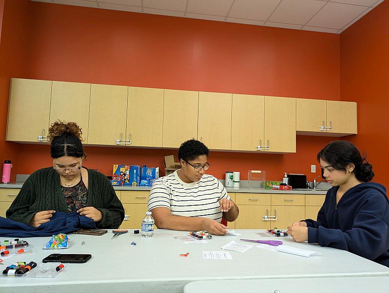 A person sitting at a table models stitches for another person who looks on attentatively.